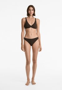 Barna bikini szett, amely csavart, V nyakú bralette topból áll, széles pántokkal, és illeszkedő, alacsony derekú alsóból készült, texturált anyagból.