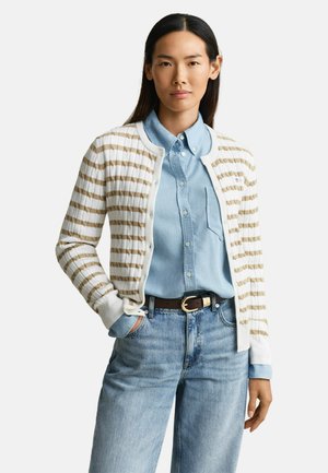 Jonge vrouw die een lichtblauw overhemd draagt, een wit-beige gestreepte cardigan, blauwe spijkerbroek en een zwarte riem, staand met één hand in de zak.