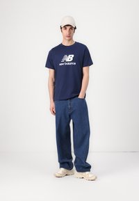 Sötétkék pamut póló, fehér "New Balance" logóval, lazán illeszkedő kék farmernadrággal és világos bézs sportcipővel párosítva. Világosszínű sapka viselve.