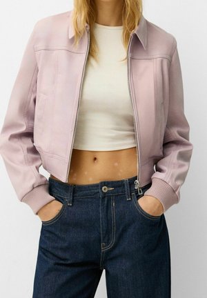 Vrouw draagt een lichtroze jas met rits over een witte cropped top en hoog gesloten donkerblauwe spijkerbroek met handen in de zakken.