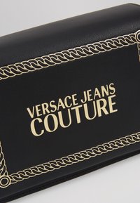 Portafoglio in pelle nera con bordo a catena dorata e logo "Versace Jeans Couture" stampato in oro sul davanti.