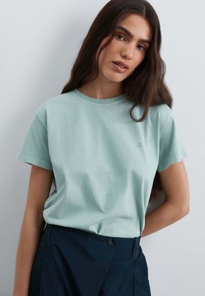 Joven mujer con pelo largo y oscuro, que lleva una camiseta de manga corta de color verde claro y pantalones oscuros de talle alto, de pie frente a una pared clara y lisa.