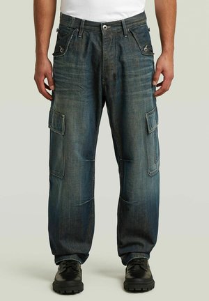 Mann trägt lockere dunkelblaue Denim-Cargohosen mit Klapptaschen und schwarze Schnürschuhe, steht vor hellem Hintergrund.