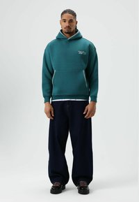 Turquoise hoodie met voorzak en ritssluiting accenten, gecombineerd met oversized donkerblauwe jeans. Simpele logodetail op de borst. Soepele stoftextuur.