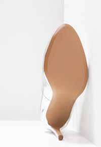 Semelle d'une chaussure à talon haut argenté brillant avec un bout pointu et un talon fin, reposant contre une surface blanche d'angle.