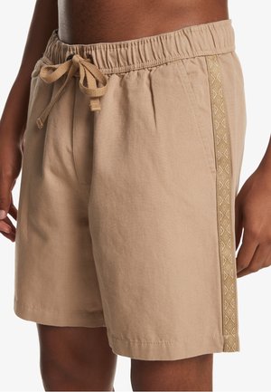 Beige elastic waist shorts with a drawstring and patterned side stripe worn by a person with dark skin.