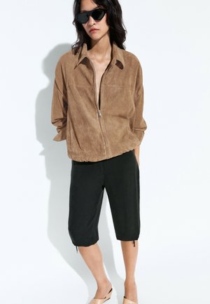Femme portant des lunettes de soleil noires, une veste zippée en daim marron, un short noir longueur genou et des chaussures beige, debout sur fond blanc.