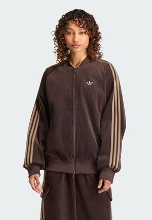 Giovane donna con capelli ricci indossa una giacca marrone Adidas con zip e strisce beige sulle maniche e pantaloni abbinati con tasche.