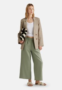 Blazer de cuadros beige sobre una camiseta blanca, combinado con pantalones verdes holgados y sandalias de plataforma color crema, sosteniendo un bolso negro y blanco con estampado.