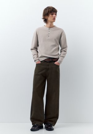 Mladá osoba stojaca s rukami v kapsách, oblečená v béžovej dlhorukávej henley košeli, tmavých širokých nohaviciach, hnedom opasku a čiernych topánkach.