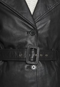 Black leather jacket with large lapels, a belted waist, and a button closure. Features textured material and silver accent hardware.