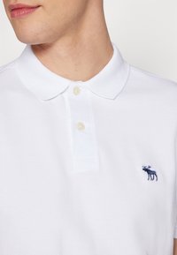 Muž oblečený v bílém polo tričku se dvěma knoflíky a malým tmavě modrým logem losa na levé straně hrudi.