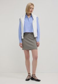 Giovane donna con una camicia button-up azzurro chiaro, felpa bianca sulle spalle, gonna a quadretti e ballerine nere con cinturini.