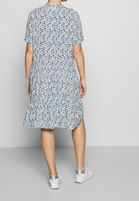 Blumenkleid mit kurzen Ärmeln, gestuftem Saum, hellblauem Hintergrund und dunklen Blumenmustern. Dazu weiße Sneaker.