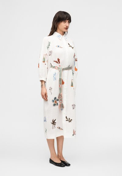 Γυναίκα που φοράει λευκό μίντι φόρεμα με κουμπιά, πολύχρωμη αφηρημένη κέντη μα και ριγωτή ζώνη, συνδυασμένο με μαύρα φλατ παπούτσια, σε προφίλ όψη.