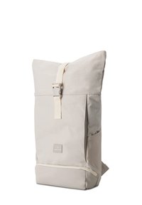 Beige színű, felhajtható hátizsák elülső csattal, oldalsó zsebekkel és kis logóval az elülső részen, fehér háttér előtt függőlegesen állva.