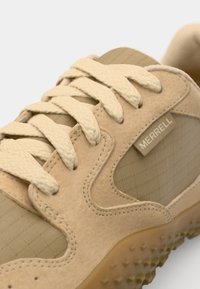 Beige athletic shoe featuring suede and textured mesh materials, round laces, and a rubber outsole with small raised patterns. Brand label visible.