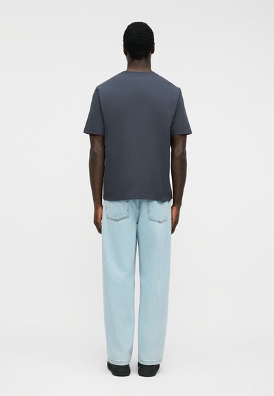 Άνδρας στέκεται γυρισμένος με την πλάτη, φορώντας σκούρο γκρι κοντομάνικο πουκάμισο, ανοιχτόχρωμο μπλε τζιν κανονικής γραμμής και μαύρα παπούτσια, μπροστά σε απλό λευκό φόντο.