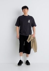 Άνδρας με μαύρο φαρδύ μπλουζάκι, μαύρα σορτς μέχρι το γόνατο, λευκές κάλτσες, μαύρα παπούτσια, κρατώντας ένα μπεζ σακάκι σε λευκό φόντο.