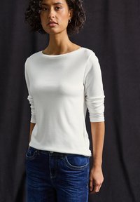 Haut blanc à manches longues avec col rond et coupe ajustée, associé à un jean bleu foncé. Texture lisse avec des détails subtils.