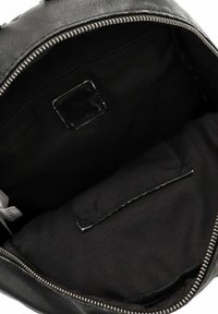 Intérieur d'un sac à dos en cuir noir, comprenant une poche zippée et une poche plate contre une doublure en tissu noir mat. Fermetures éclair en métal argenté.