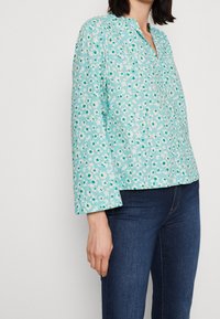 Turkos blommig blus med mandarin-krage, långa ärmar, som har ett mönster av vita och gröna blommor på ett mjukt, strukturerat tyg.