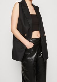 Blazer noir sans manches à texture lisse, avec des boutons dorés et des revers nets, associé à un crop top noir et un pantalon en cuir brillant.