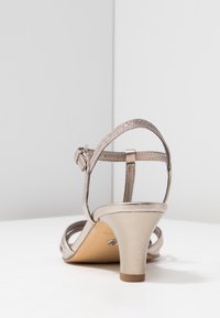 Sandale ouverte en beige métallique avec un talon conique de hauteur moyenne et une bride de cheville réglable sur une surface blanche contre un arrière-plan gris clair.