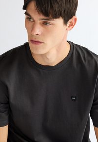 Jeune homme aux cheveux courts et foncés portant un t-shirt noir simple Wrangler, regardant vers sa gauche sur un fond clair.