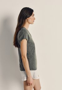 Jauna moteris su ilgais rudais plaukais stovi šone, dėvi pilką marškinėlį ir šviesių spalvų trumpikes, fone paprasta siena.