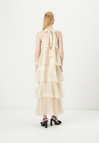 Tiered cream silk dress with a halter neck and bow detail at the back, featuring layered ruffles and a flowing silhouette. Black heeled sandals.