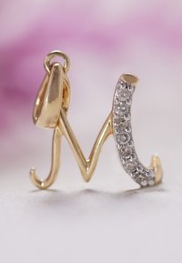 Gouden letter "M" hanger met een gepolijste afwerking en een rij kleine, heldere rhinestones aan de ene kant. Gebogen en elegante vormgeving.