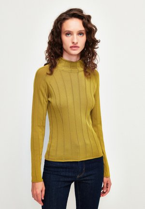 Mosterdgele lange mouw top met hoge halslijn, voorzien van ribbeltextuur en verticale strepen, gecombineerd met donkerblauwe jeans.