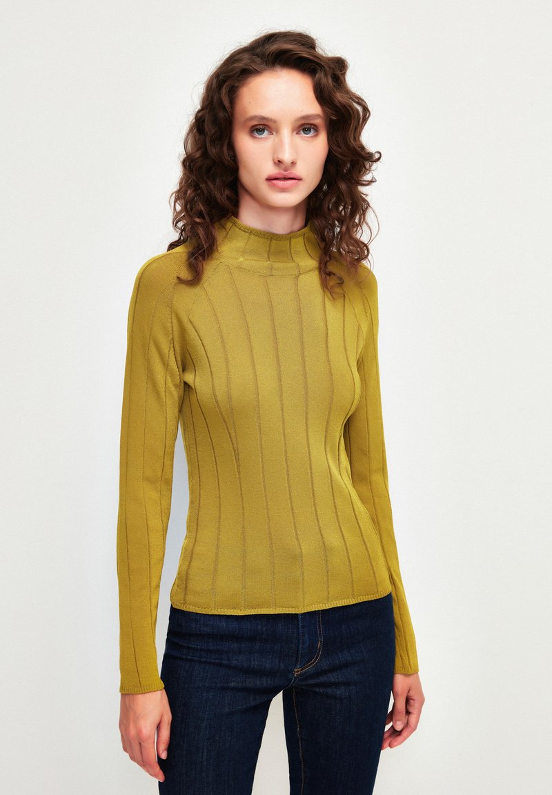 Senfgelbes Langarmshirt mit hohem Kragen, das über eine gerippte Textur und vertikale Streifen verfügt, kombiniert mit dunkelblauen Jeans.