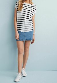 Kvinde iført en hvid og sort stribet udenærmet top, denim mini nederdel, hvide sneakers, stående mod en lyseblå væg.