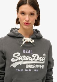 Felpa grigia in cotone con cappuccio regolabile, caratterizzata da un audace logo e testo "SuperDry" in bianco e viola, e una tasca a marsupio davanti.