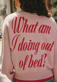 Růžový pletený svetr s uvolněným střihem. Na zadní straně se nachází velký červený text "Co to dělám venku z postele?". Měkká textura.