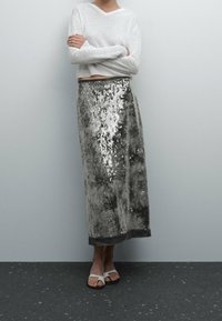 Jupe longue en sequins gris avec une surface texturée, associée à un pull blanc court et des sandales blanches plates. Design longueur sol.