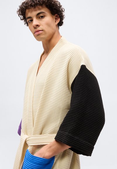 Άνδρας που φορά ρόμπα με υφή βάφλας σε κρεμχρώμα στο σώμα, με μαύρο δεξί μανίκι, μωβ αριστερό μανσέτι και μπλε τσέπη, στέκεται μπροστά σε λευκό φόντο.