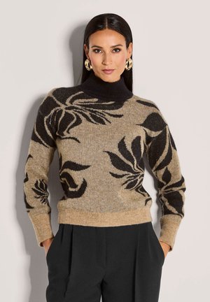 Frau trägt einen beige-schwarzen, floral gemusterten Pullover mit einem schwarzen Rollkragen und schwarzen Hosen, die Hände in den Taschen, vor einem schlichten Hintergrund.