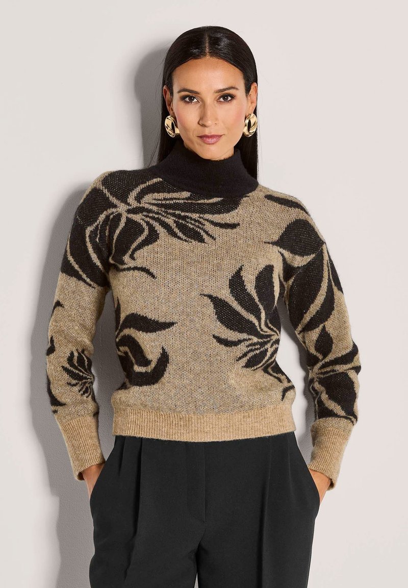 Frau trägt einen beige-schwarzen, floral gemusterten Pullover mit einem schwarzen Rollkragen und schwarzen Hosen, die Hände in den Taschen, vor einem schlichten Hintergrund.