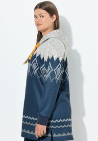 Hoodie lungo blu marina con cappuccio grigio, caratterizzato da un motivo a diamante bianco e grigio. Tessuto morbido, polsini a coste e spacchi laterali.