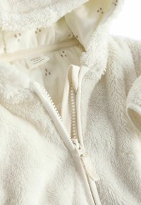 Cremefarbene, flauschige Baby-Einteiler mit einem Reißverschluss, weichem Material, einer Kapuze und einem gemusterten Innenfutter mit kleinen braunen Punkten.