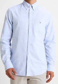 Camisa de botones azul claro hecha de algodón, con cuello, mangas largas y un bolsillo en el pecho con un emblema. Textura suave, diseño clásico.