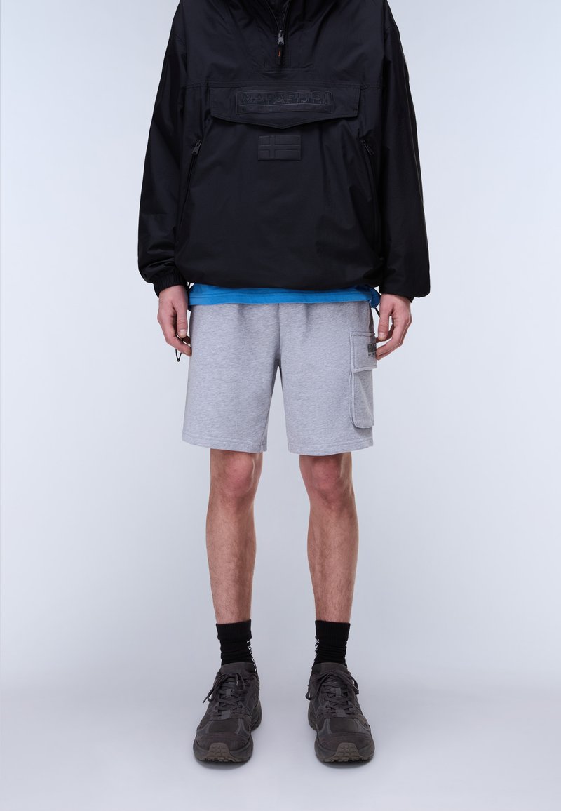 Άτομο που φοράει μαύρο μπουφάν, μπλε πουκάμισο, γκρι σορτς τύπου cargo, μαύρες κάλτσες και μαύρα αθλητικά παπούτσια, στέκεται μπροστά σε ένα απλό φωτεινό φόντο.