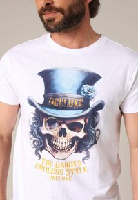 Witte katoenen T-shirt met een grafische print van een schedel in een blauwe hoge hoed met een roos en gouden tekst met de woorden "DE DANDY'S EINDLOZE STIJL."
