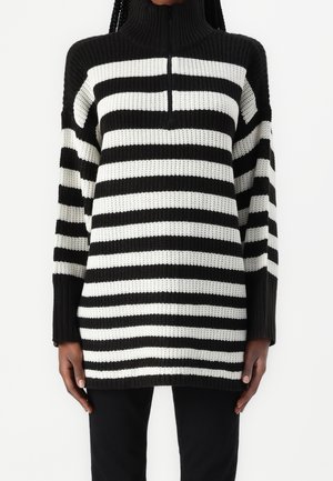 Osoba ubrana w sweter dzianinowy z poziomymi czarno-białymi paskami, z wysokim kołnierzem i półzamkiem, w połączeniu z czarnymi spodniami.