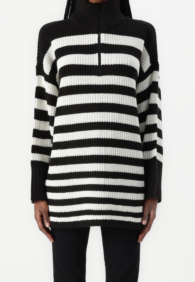 Personne portant un pull en tricot à rayures horizontales noires et blanches avec un col montant et une demi-fermeture éclair, associé à un pantalon noir.
