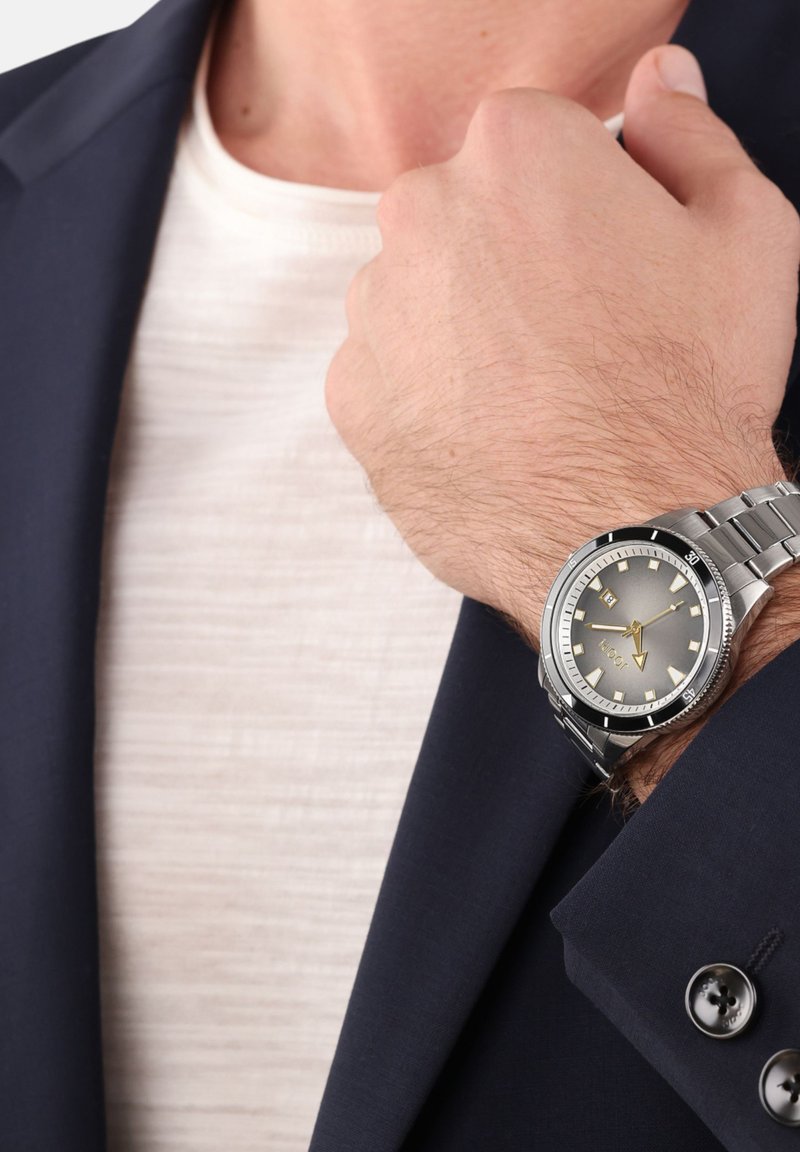 Zilveren metalen horloge met een grijze wijzerplaat, luminescente wijzers en markeringen. Draagbaar om een pols, met een marineblauwe blazer en een crème shirt zichtbaar.