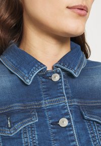 Jeansjacke mit klassischem Kragen und silberner Druckknopfverschluss. Der Stoff ist mittelblau mit leichter Ausbleichung und kontrastierenden Nähten.